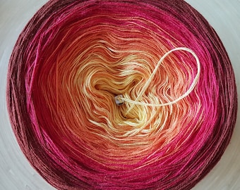 May include: A vibrant gradient yarn cake in shades of burgundy, fuchsia, orange, and gold. The yarn is wound in a circular pattern, with a loop of thread extending from the center. A small, square wooden bead is attached to the thread.