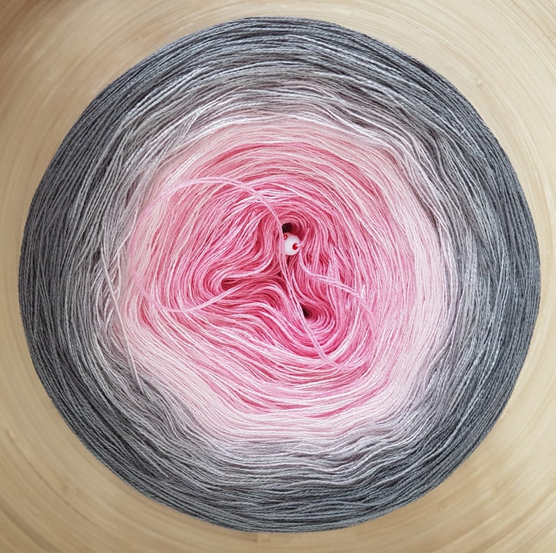 May include: A ball of yarn in a gradient of colours, from dark grey to light pink in the centre. The yarn is on a wooden surface. The yarn ball is a craft supply for knitting or crochet.