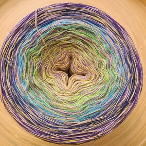 May include: A vibrant, multi-colored yarn cake with a gradient effect. The yarn transitions from purple to blue, teal, green, yellow, and back to purple. The yarn is arranged in a circular pattern, with a central opening.