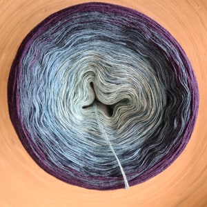 May include: A round ball of yarn with a gradient of colors, transitioning from deep purple to light blue and green. The yarn is tightly wound, with a single strand extending from the center. The yarn is likely for knitting or crocheting.