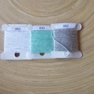 Op de afbeelding: Drie spoelen garen in wit, teal en grijs, gelabeld met 001, 631 en 002. Het garen is gewikkeld op witte plastic spoelen. De achtergrond is een lichtbruin houten oppervlak.