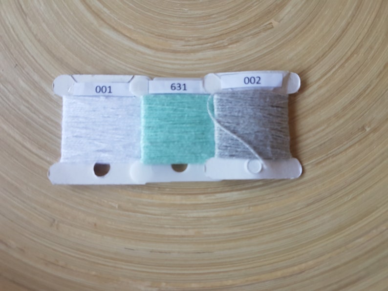 Op de afbeelding: Drie spoelen garen in wit, teal en grijs, gelabeld met 001, 631 en 002. Het garen is gewikkeld op witte plastic spoelen. De achtergrond is een lichtbruin houten oppervlak.