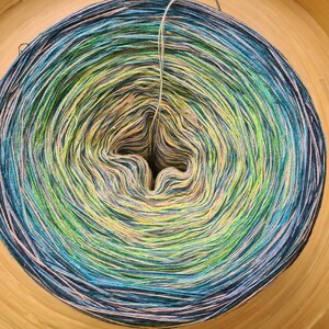 May include: A large, colorful yarn cake with a gradient of colors, including blues, greens, and yellows. The yarn is tightly wound, creating a spiral pattern. A single strand of yarn extends from the center. The yarn is set in a wooden bowl.