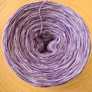 May include: A tightly wound ball of yarn in shades of purple and white, with a single strand extending from the center. The yarn is arranged in concentric circles, creating a spiral pattern. The yarn is on a light brown wooden surface.