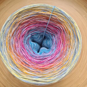 May include: A tightly wound ball of yarn with a gradient of colors, including blue, pink, yellow, and white. The yarn is centered with a deep blue hue, transitioning outwards through vibrant shades. A single strand of yarn extends from the center.