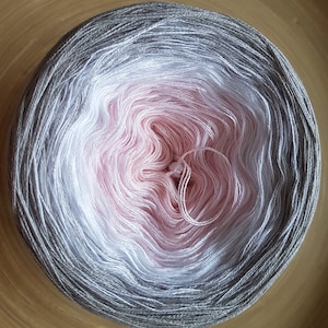 Può includere: Un gomitolo di filato con una sfumatura di colore, che passa dal grigio al bianco e poi al rosa al centro. Il filato è ben avvolto, con fili visibili al centro. Il filato è su una superficie di legno.