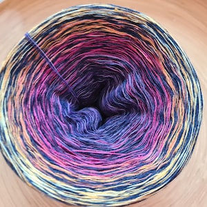 May include: A tightly wound ball of yarn with a gradient of colors. The yarn transitions from yellow to orange, then to pink, purple, and finally to a deep navy blue. A single strand of the purple yarn extends from the center.