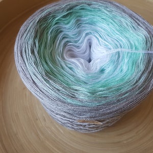 Op de afbeelding: Een bol garen in tinten grijs, wit en blauwgroen, rustend in een houten kom. Het garen heeft een gradi&euml;nteffect, dat overgaat van donkerder grijs naar lichtere tinten. Een paar strengen garen steken uit de bol.