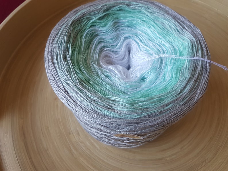 Op de afbeelding: Een bol garen in tinten grijs, wit en blauwgroen, rustend in een houten kom. Het garen heeft een gradi&euml;nteffect, dat overgaat van donkerder grijs naar lichtere tinten. Een paar strengen garen steken uit de bol.