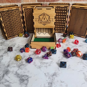 May include: A wooden dice tower with a green felt bottom and a clear plastic tray. The tower has a sign that says "Crit Happens" and a die with a 1 on it. There are many colorful dice scattered around the tower on a white marble surface.