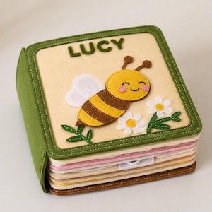 May include: A square felt quiet book with a cream-colored cover featuring the name LUCY in dark green letters. The cover displays a yellow and brown bee with white wings, two white daisies with yellow centers, and green stitched leaves.