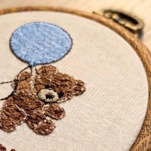 May include: Embroidered teddy bear holding a blue balloon. The brown bear is stitched on a beige fabric, set within a wooden embroidery hoop. The design features detailed stitching, creating a textured appearance. The overall theme is a handmade craft.