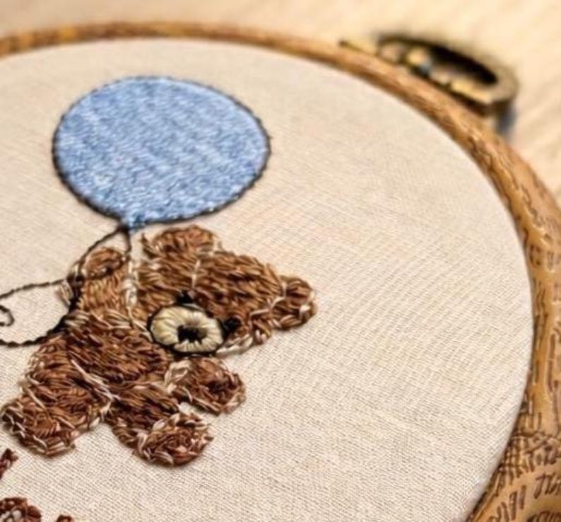 May include: Embroidered teddy bear holding a blue balloon. The brown bear is stitched on a beige fabric, set within a wooden embroidery hoop. The design features detailed stitching, creating a textured appearance. The overall theme is a handmade craft.