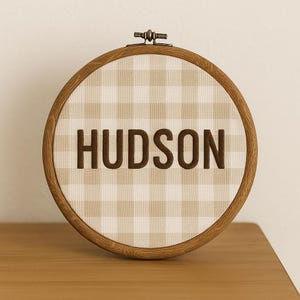 May include: A wooden embroidery hoop with a tan and white gingham fabric. The word "HUDSON" is embroidered in brown thread across the center. The hoop is resting on a wooden surface.