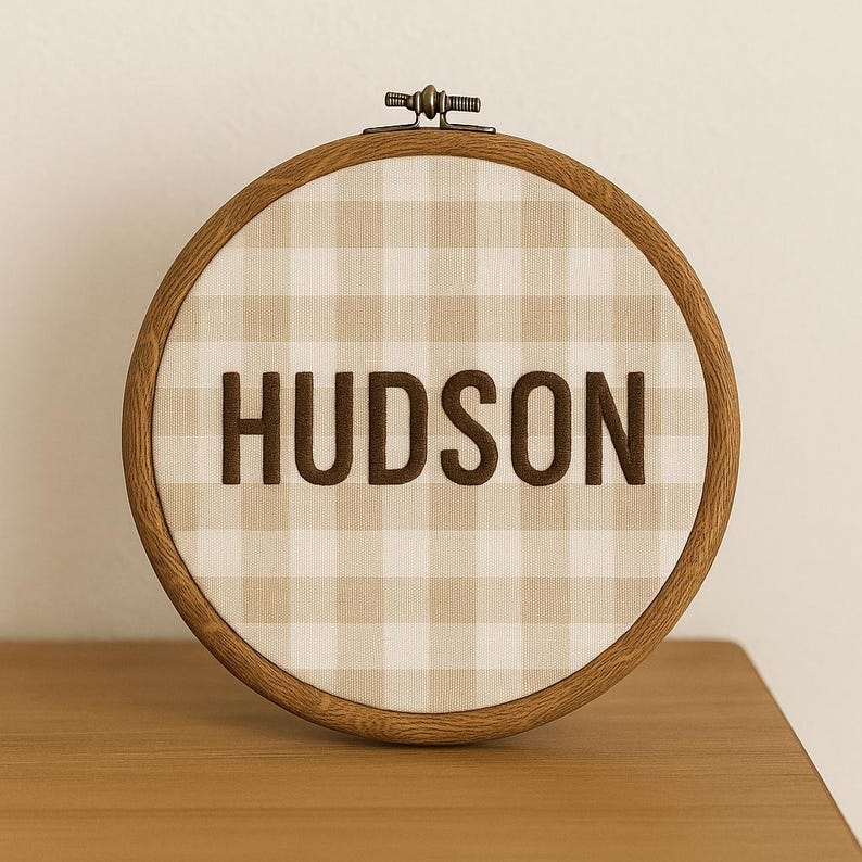 May include: A wooden embroidery hoop with a tan and white gingham fabric. The word "HUDSON" is embroidered in brown thread across the center. The hoop is resting on a wooden surface.