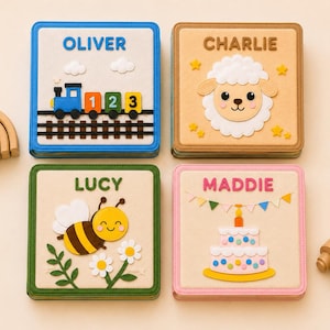 May include: Four square fabric books with embroidered names and felt appliques. The top left book features a blue train with the name OLIVER. The top right shows a white sheep and the name CHARLIE. The bottom left displays a bee and the name LUCY. The bottom right shows a cake and the name MADDIE.