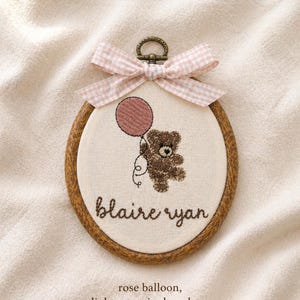 May include: Embroidered hoop art featuring a brown teddy bear holding a rose-colored balloon. The bear is suspended in the air. The name "blaire ryan" is stitched below the bear. A light rose gingham bow tops the hoop.