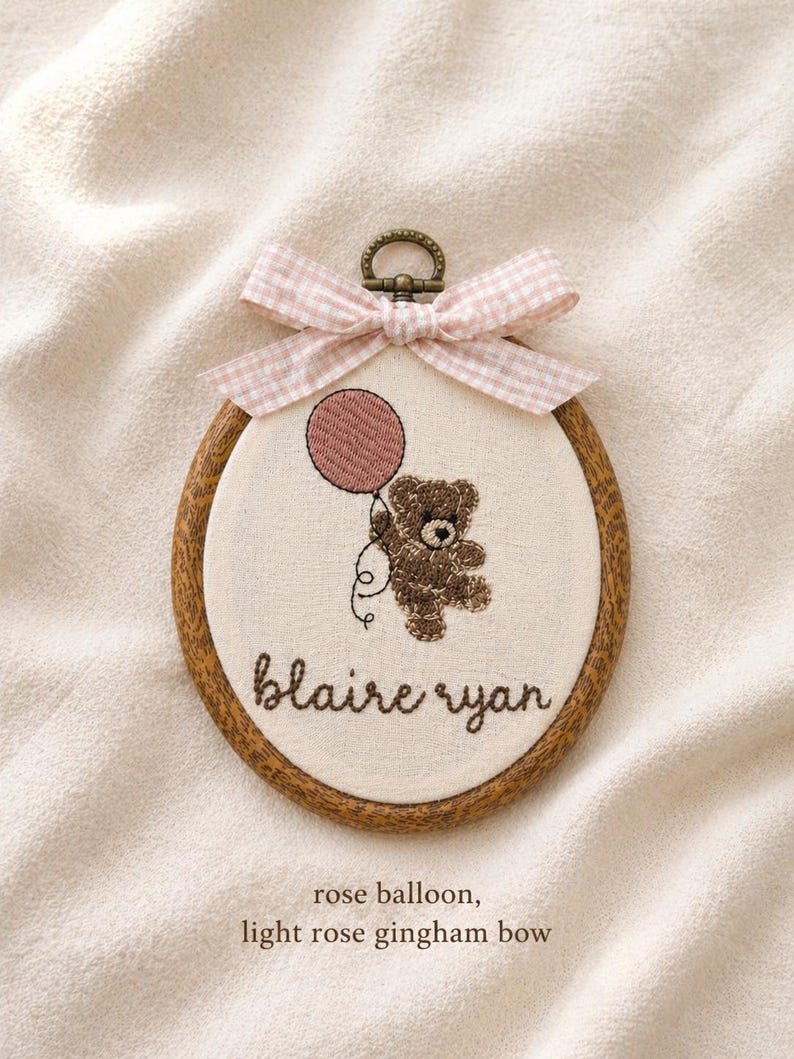 May include: Embroidered hoop art featuring a brown teddy bear holding a rose-colored balloon. The bear is suspended in the air. The name "blaire ryan" is stitched below the bear. A light rose gingham bow tops the hoop.