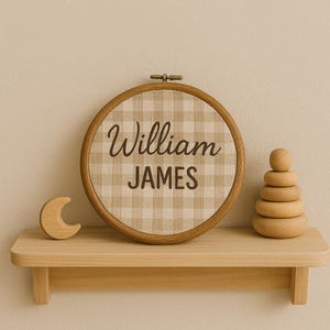May include: A wooden shelf displays a round embroidery hoop with the name "William JAMES" on a tan and white checkered fabric. A wooden crescent moon and a wooden stacking toy are also on the shelf.