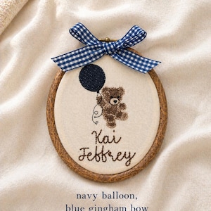 May include: Embroidered hoop art featuring a brown teddy bear holding a navy blue balloon. The text "Kai Jeffrey" is stitched below the bear. A blue and white gingham bow adorns the top of the wooden hoop.