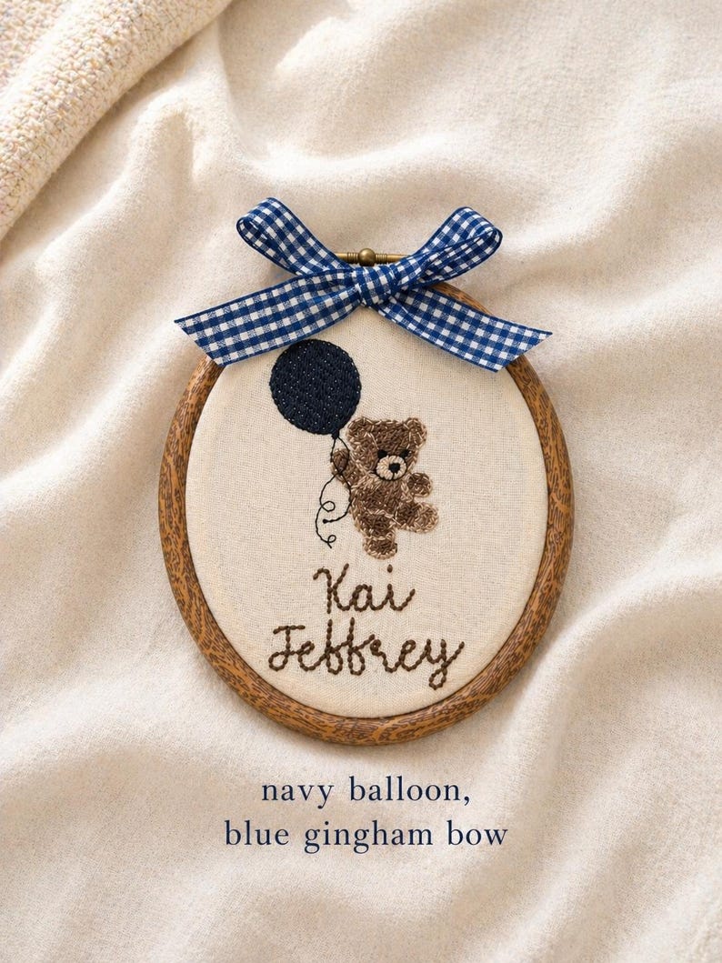 May include: Embroidered hoop art featuring a brown teddy bear holding a navy blue balloon. The text "Kai Jeffrey" is stitched below the bear. A blue and white gingham bow adorns the top of the wooden hoop.
