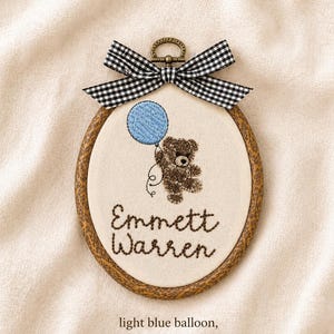 May include: Embroidered oval hoop art featuring a brown teddy bear holding a light blue balloon. The text "Emmett Warren" is stitched below the bear. A black and white gingham bow tops the hoop.