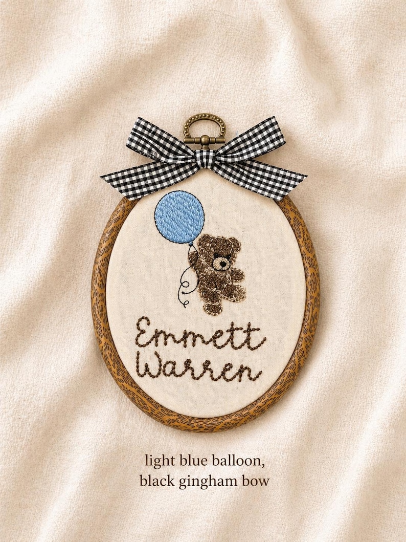 May include: Embroidered oval hoop art featuring a brown teddy bear holding a light blue balloon. The text "Emmett Warren" is stitched below the bear. A black and white gingham bow tops the hoop.