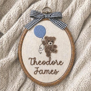 May include: An embroidered oval hoop art piece featuring a brown teddy bear holding a blue balloon. The text "Theodore James" is stitched in brown thread. A black and white checkered bow tops the wooden frame.