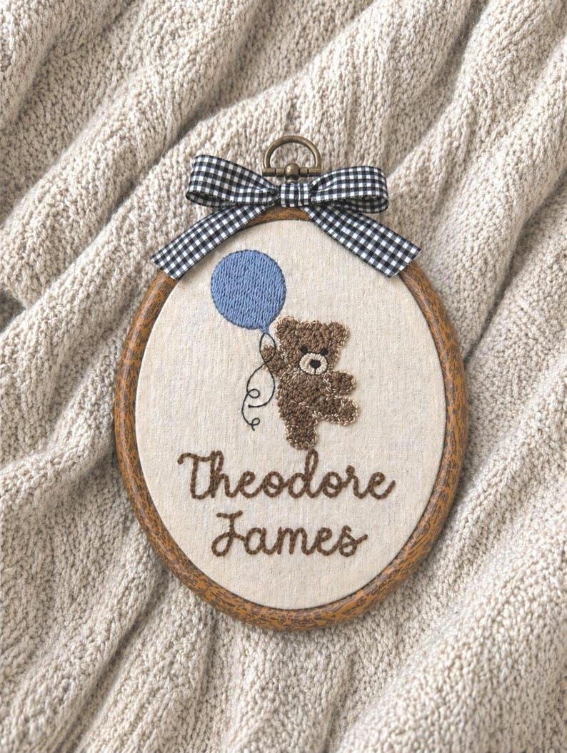 May include: An embroidered oval hoop art piece featuring a brown teddy bear holding a blue balloon. The text "Theodore James" is stitched in brown thread. A black and white checkered bow tops the wooden frame.