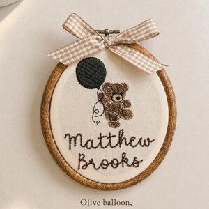 May include: Embroidered hoop art featuring a brown teddy bear holding an olive green balloon. The name "Matthew Brooks" is stitched below the bear. A beige gingham bow adorns the top of the wooden hoop. The text "Olive balloon, beige gingham bow" is at the bottom.
