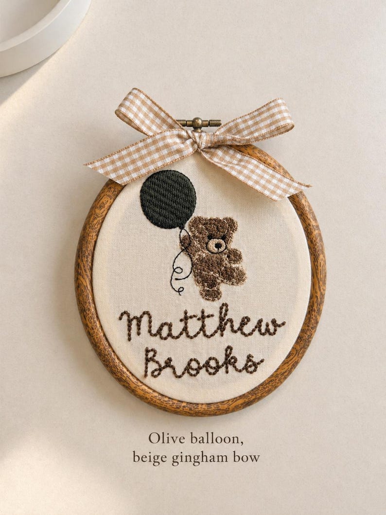 May include: Embroidered hoop art featuring a brown teddy bear holding an olive green balloon. The name "Matthew Brooks" is stitched below the bear. A beige gingham bow adorns the top of the wooden hoop. The text "Olive balloon, beige gingham bow" is at the bottom.