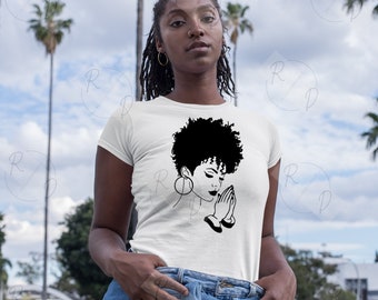 Op de afbeelding: Een wit T-shirt met een zwart-witte illustratie van een vrouw met haar handen gevouwen in gebed. De vrouw heeft kort, krullend haar en draagt een grote oorring.