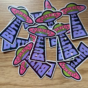 May include: A pile of colorful, holographic stickers featuring a cartoon UFO with the word "BUTTS" written on it. The stickers have a glittery finish and are perfect for adding a fun and quirky touch to any surface.