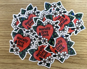 May include: A stack of red heart-shaped stickers with the text "Morally Grey Men" in white lettering. The stickers are surrounded by white flowers and green leaves.