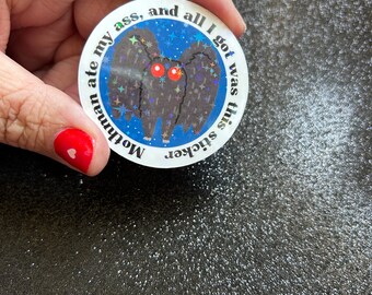 Puede incluir: Una pegatina redonda con un diseño de Mothman de dibujos animados. El Mothman tiene ojos rojos y un cuerpo oscuro y texturizado, sobre un fondo azul estrellado. El texto dice "Mothman ate my ass, and all I got was this sticker".