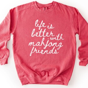 May include: A watermelon-colored sweatshirt with the phrase "life is better with mahjong friends" in white script. The long-sleeved crewneck shirt has a relaxed fit and is made of a soft material. The bottom right corner of the shirt has the word "watermelon".