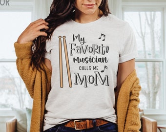 Pode incluir: Uma camiseta branca com um gráfico preto que diz "My Favorite Musician Calls Me Mom" com um par de baquetas e notas musicais.