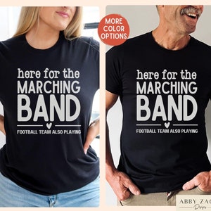 May include: Two black t-shirts with white text that reads "here for the marching band" and "football team also playing".
