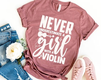 May include: A heather mauve t-shirt with the text "Never underestimate a girl with a violin" in white. The shirt is shown on a white background with a pair of pink sneakers, a pair of blue jeans, and a white flower.