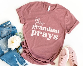 May include: A heather mauve t-shirt with the text "This grandma prays" in white. The shirt is shown on a white background with a pair of pink sneakers, a white flower, and a pair of blue jeans.