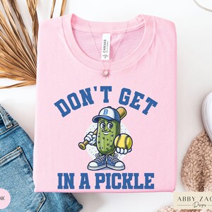 Puede incluir: Camiseta rosa con un pepinillo de dibujos animados que lleva una gorra de b&eacute;isbol y sostiene un bate y una pelota de b&eacute;isbol. El texto "Don't Get In A Pickle" est&aacute; impreso en la camiseta.