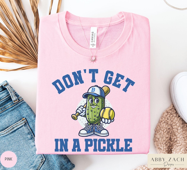 Puede incluir: Camiseta rosa con un pepinillo de dibujos animados que lleva una gorra de b&eacute;isbol y sostiene un bate y una pelota de b&eacute;isbol. El texto "Don't Get In A Pickle" est&aacute; impreso en la camiseta.