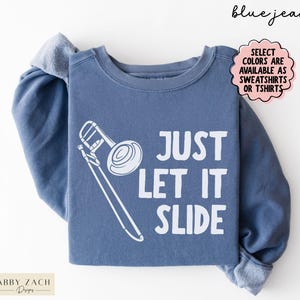 May include: A blue jean colored crewneck sweatshirt featuring a white graphic of a trombone and the text "JUST LET IT SLIDE". The image includes text in the top right corner stating "blue jean" and a pink cloud shape with text about color availability.