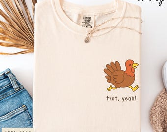 May include: Ivory-colored Comfort Colors t-shirt with a cartoon turkey graphic. The turkey is brown, red, and yellow, with the text "trot, yeah!" below. The shirt has a crew neck and a small tag.