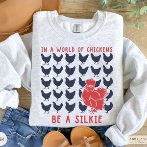 May include: A gray sweatshirt with the text "In a world of chickens, be a silkie" printed on it. The text is in black and red. There are multiple black silhouettes of chickens surrounding the text.
