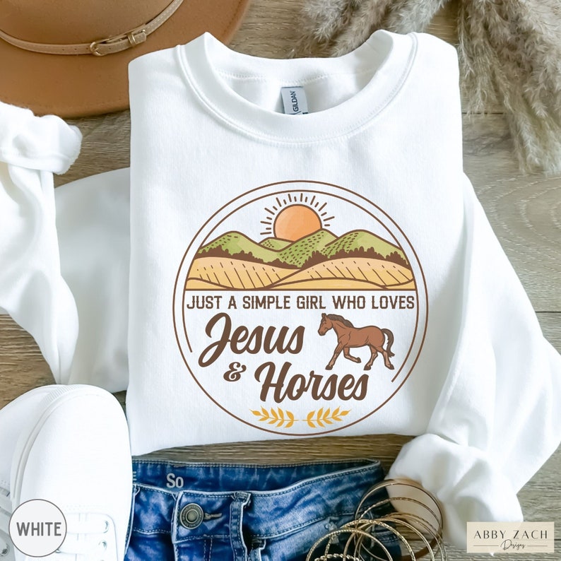 K&ouml;nnte beinhalten: Wei&szlig;er Pullover mit einem Grafikdesign eines Sonnenuntergangs &uuml;ber einem Feld mit dem Text "Just a simple girl who loves Jesus & Horses".