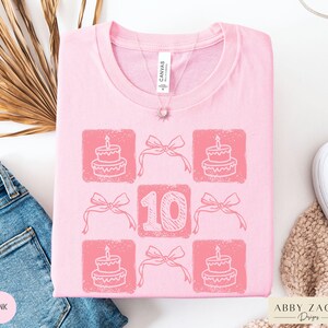Peut inclure: T-shirt rose avec un motif graphique d'un g&acirc;teau d'anniversaire, des n&oelig;uds et le chiffre 10. Le texte "Abby Zach Designs" est dans le coin inf&eacute;rieur droit.