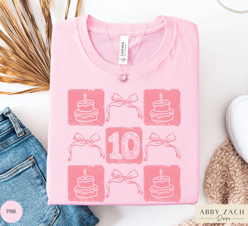 Peut inclure: T-shirt rose avec un motif graphique d'un g&acirc;teau d'anniversaire, des n&oelig;uds et le chiffre 10. Le texte "Abby Zach Designs" est dans le coin inf&eacute;rieur droit.