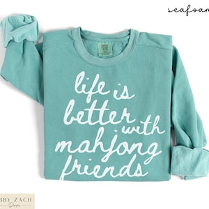 May include: Seafoam green sweatshirt with the text "life is better with mahjong friends" in white script. The long-sleeved shirt has a crew neck and rolled cuffs. The brand name "Abby Zach Designs" is at the bottom left.