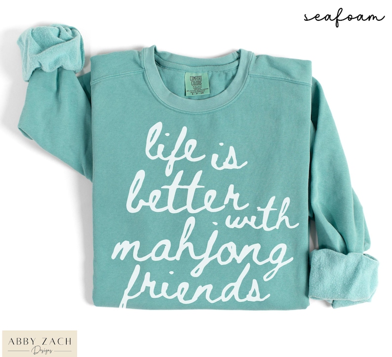 May include: Seafoam green sweatshirt with the text "life is better with mahjong friends" in white script. The long-sleeved shirt has a crew neck and rolled cuffs. The brand name "Abby Zach Designs" is at the bottom left.